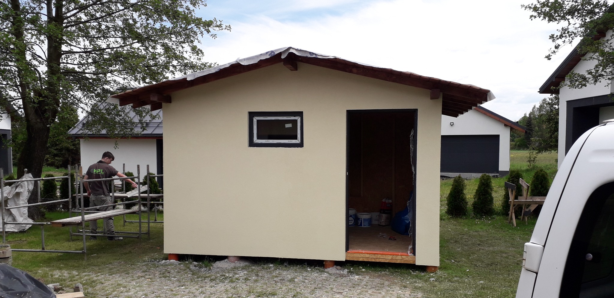 Nowo wybudowany, otynkowany domek gospodarczy z oknem i otwartymi drzwiami, widoczne puszki z farbą w środku, na zewnątrz pracownik na rusztowaniu, w tle inne budynki.