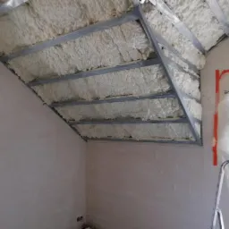 Poddasze z widoczną konstrukcją metalową wypełnioną białą pianką izolacyjną, okno zabezpieczone folią malarską, drabina oparta o ścianę.