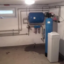 Instalacja uzdatniania wody w piwnicy: zbiornik ciśnieniowy, filtr narurowy i zmiękczacz wody, połączone rurami z zaworami, widoczne ślady wilgoci na posadzce.