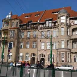 Kamienica z czerwoną dachówką i ozdobnymi wieżyczkami, widoczna zza barierki. Fasada w odcieniach beżu i brązu, z widocznymi detalami architektonicznymi i oknami.