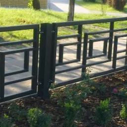 Nowoczesna, czarna metalowa furtka z geometrycznym wzorem, zamontowana przy betonowym słupku, otoczona rabatą z krzewami i korą.