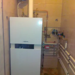 Biały piec gazowy Viessmann z niebieskim zbiornikiem wyrównawczym w kotłowni wyłożonej beżowymi płytkami, widoczne rury instalacji CO i CWU.