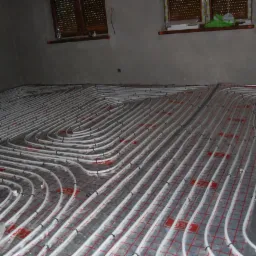 Białe rury ogrzewania podłogowego ułożone na czerwonej macie w surowym pomieszczeniu z oknami i szarymi ścianami, gotowe do zalania wylewką.