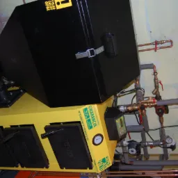 Żółto-czarny piec na paliwo stałe z czarnym zasobnikiem, widoczne przyłącza hydrauliczne i rury. Kocioł marki PER-EKO z gwarancją.