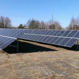 Instalacja fotowoltaiczna na dwunożnej konstrukcji, widoczna na tle betonowych płyt i drzew bez liści pod błękitnym niebem.