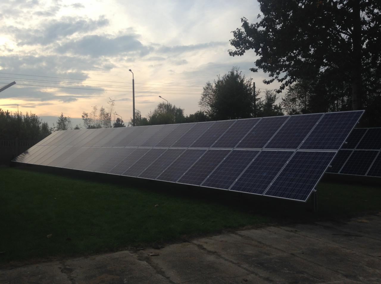 Długa instalacja fotowoltaiczna na trawniku, widok z dołu, tło: drzewa i zachmurzone niebo o zmierzchu. Panele ustawione rzędami.