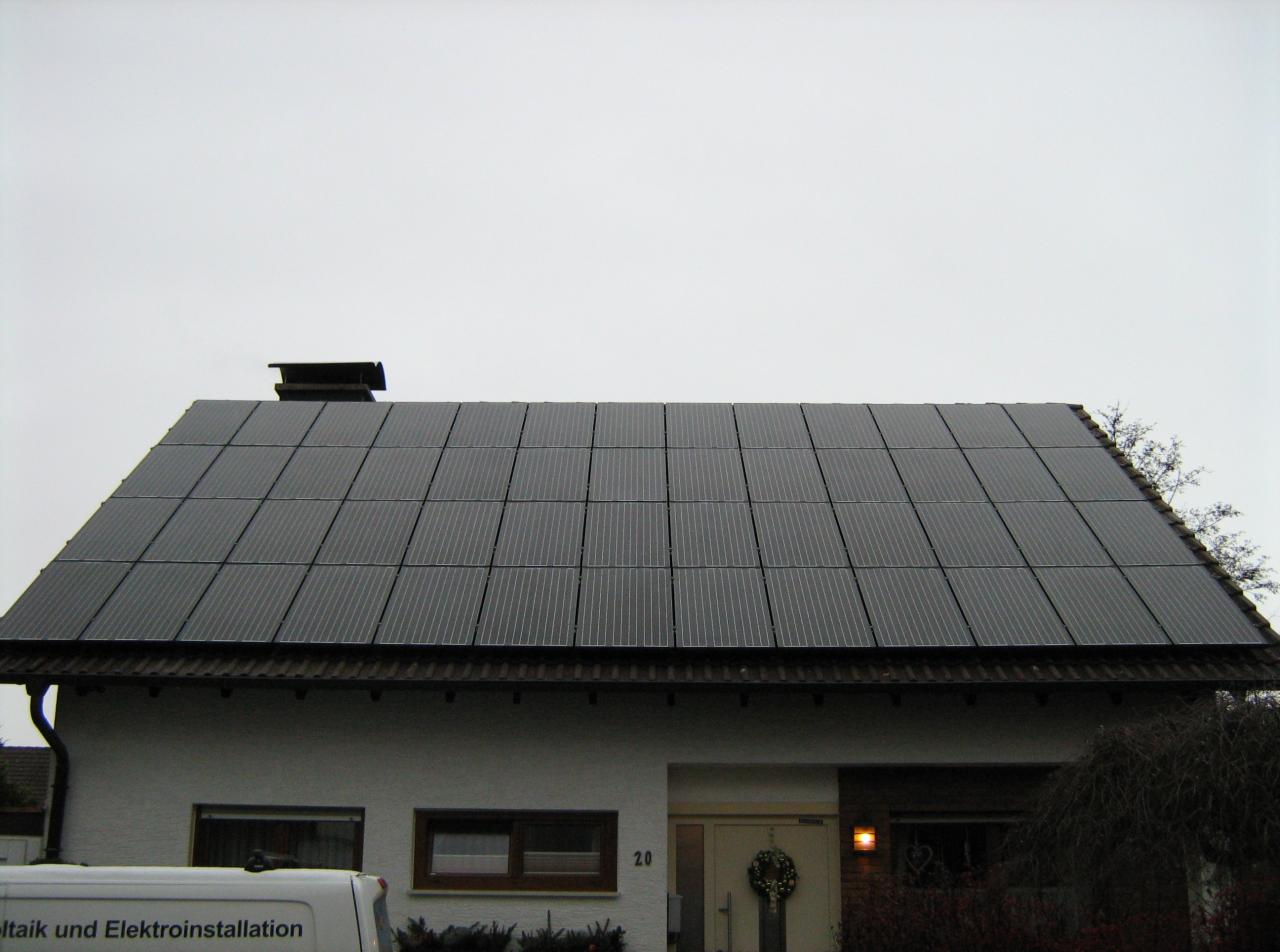 Dom jednorodzinny z panelami fotowoltaicznymi na dachu, widok z dołu. Dach pokryty panelami, komin. W tle pochmurne niebo. Częściowo widoczny samochód z napisem.