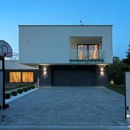 Nowoczesny dom jednorodzinny z płaskim dachem, balkonem z szklaną balustradą i koszem do koszykówki na podjeździe wyłożonym szarą kostką brukową, oświetlony zewnętrznymi lampami.