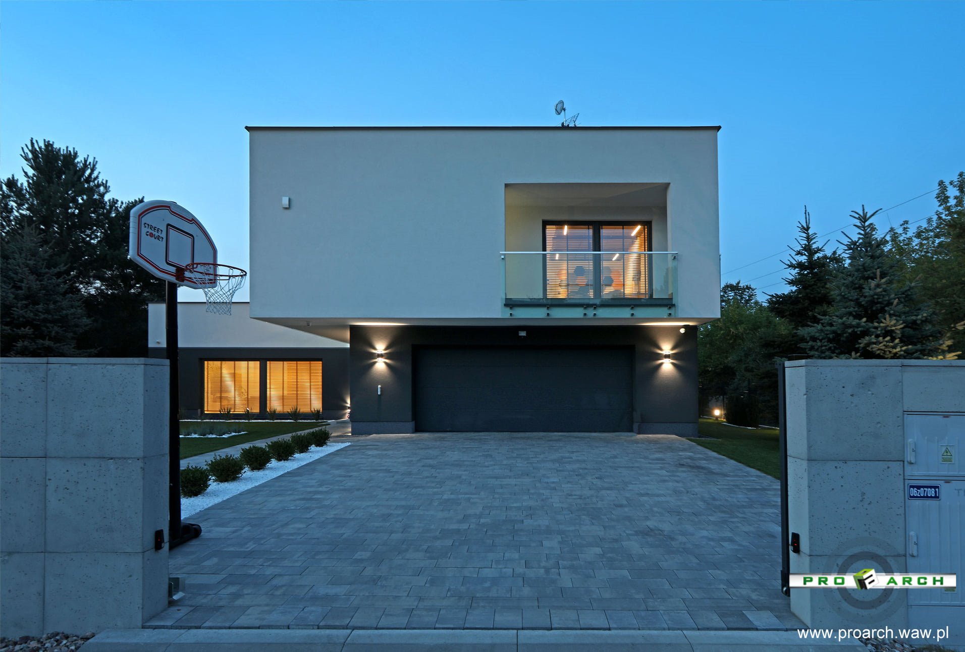Nowoczesny dom jednorodzinny z płaskim dachem, balkonem z szklaną balustradą i koszem do koszykówki na podjeździe wyłożonym szarą kostką brukową, oświetlony zewnętrznymi lampami.