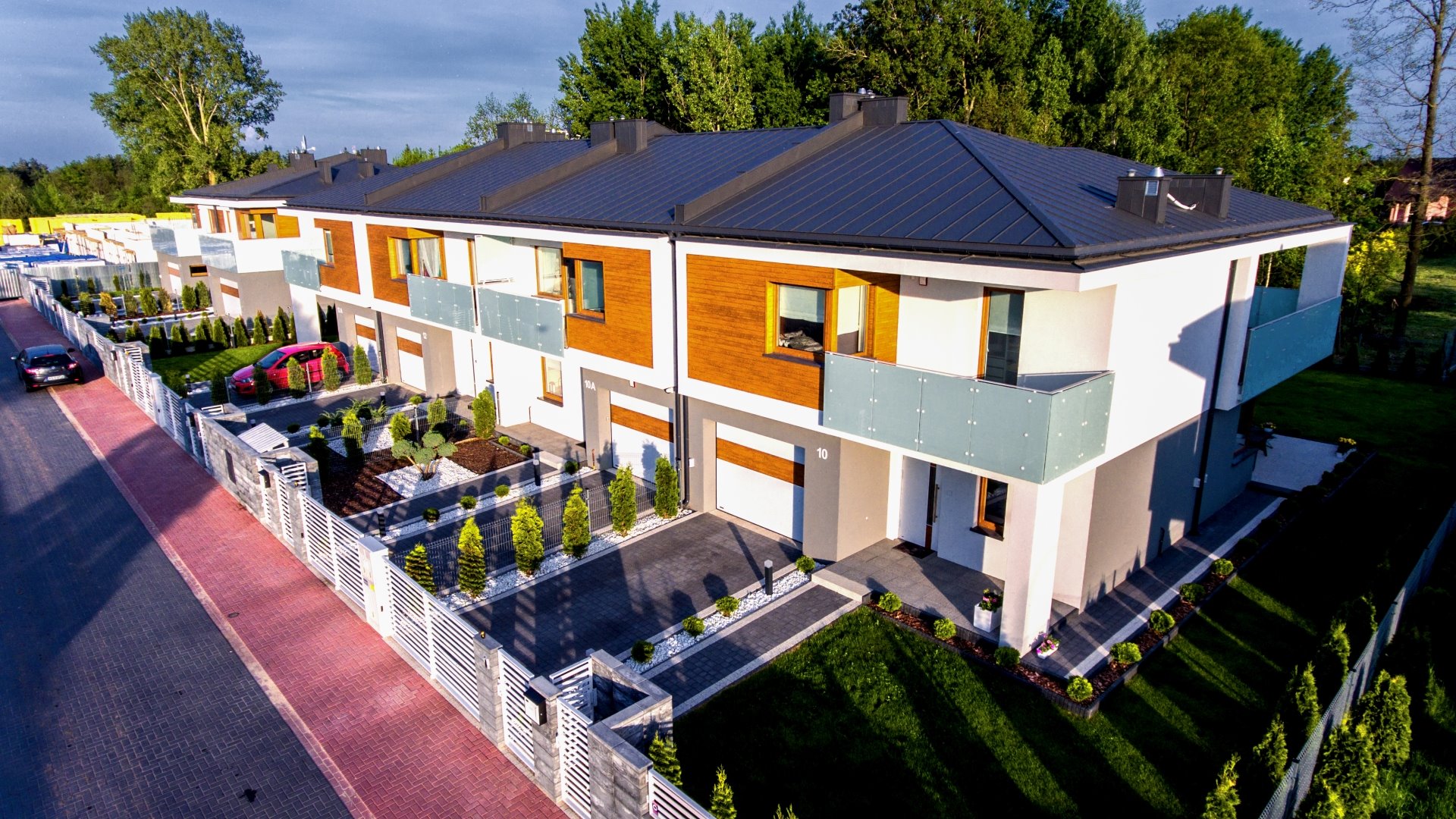 Nowoczesne szeregówki z grafitowym dachem, drewnianą elewacją i szklanymi balustradami balkonów, otoczone zadbanymi ogródkami z tujami i białym grysem, widok z lotu ptaka.