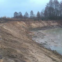Brzeg nowo wykopanego stawu z widoczną, świeżo uformowaną skarpą i wodą wypełniającą dno. W tle drzewa bez liści odbijające się w tafli wody.