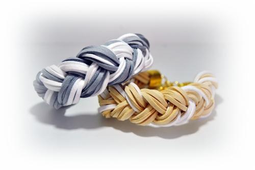 Dwa plecione bransoletki: szaro-biała i beżowo-biała, na białym tle, z widocznymi złotymi zapięciami.