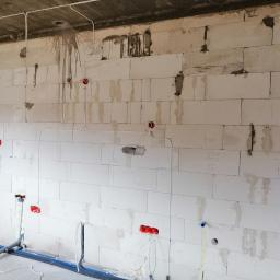 EL - KAB Usługi Elektryczne - Surowy mur z betonu komórkowego z widocznymi instalacjami elektrycznymi w postaci przewodów i puszek podtynkowych oraz instalacjami hydraulicznymi z rurami w podłodze, etap prac wykończeniowych.