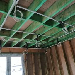 EL - KAB Usługi Elektryczne - Wnętrze w trakcie instalacji elektrycznej: białe rurki PCV mocowane do zielonej drewnianej konstrukcji stropu, z wiszącymi kablami w pętlach, widok z dołu na tle surowych ścian i okna.