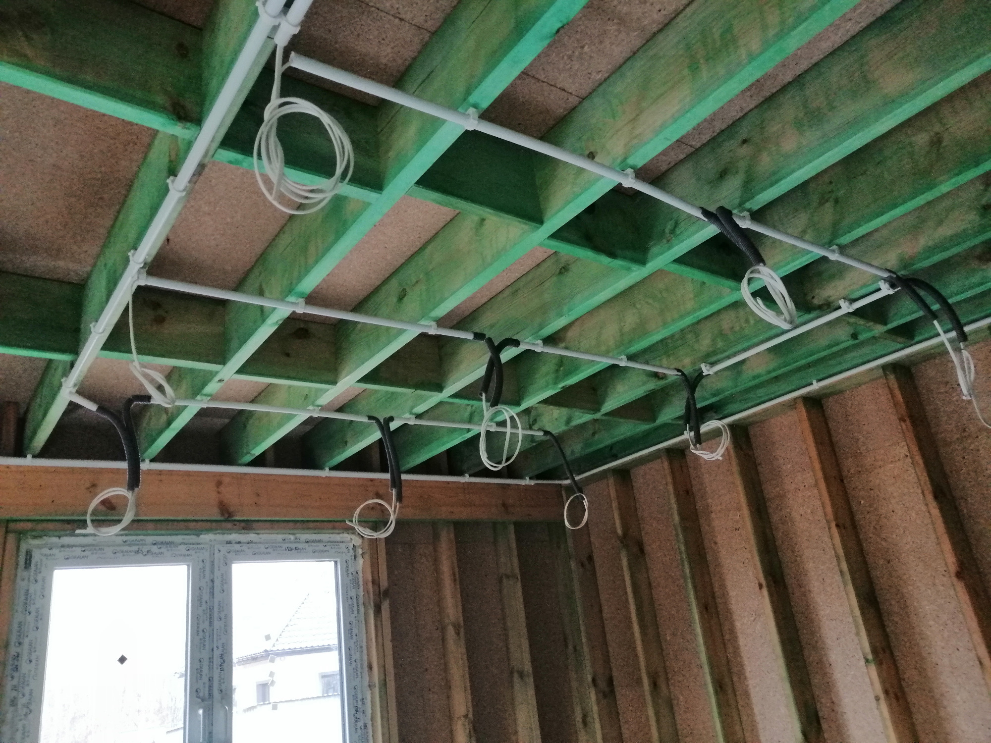 Wnętrze w trakcie instalacji elektrycznej: białe rurki PCV mocowane do zielonej drewnianej konstrukcji stropu, z wiszącymi kablami w pętlach, widok z dołu na tle surowych ścian i okna.