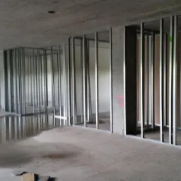 Wnętrze w trakcie budowy ścian działowych z metalowych profili i płyt kartonowo-gipsowych, widoczne kałuże na surowym betonowym podłożu.