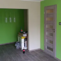 Wnętrze pokoju po odświeżeniu z zielonymi ścianami, panelami podłogowymi w odcieniach szarości i nową, drewnianą ościeżnicą drzwiową z przeszkleniami; w tle widoczne pozostałości po pracach...