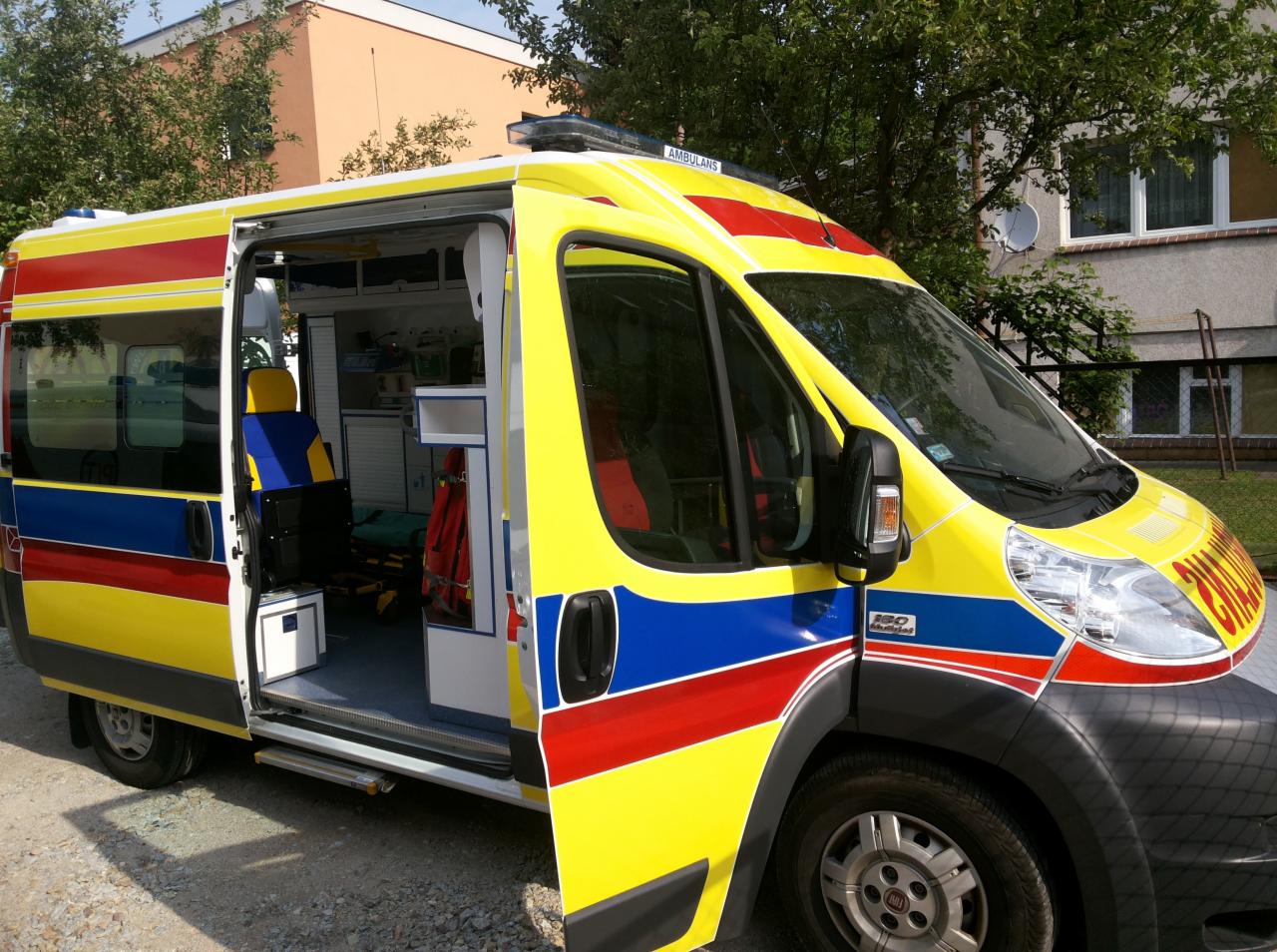 Żółto-czerwono-niebieska karetka z otwartymi drzwiami bocznymi, widoczny sprzęt medyczny wewnątrz, na zewnątrz budynki i drzewa.