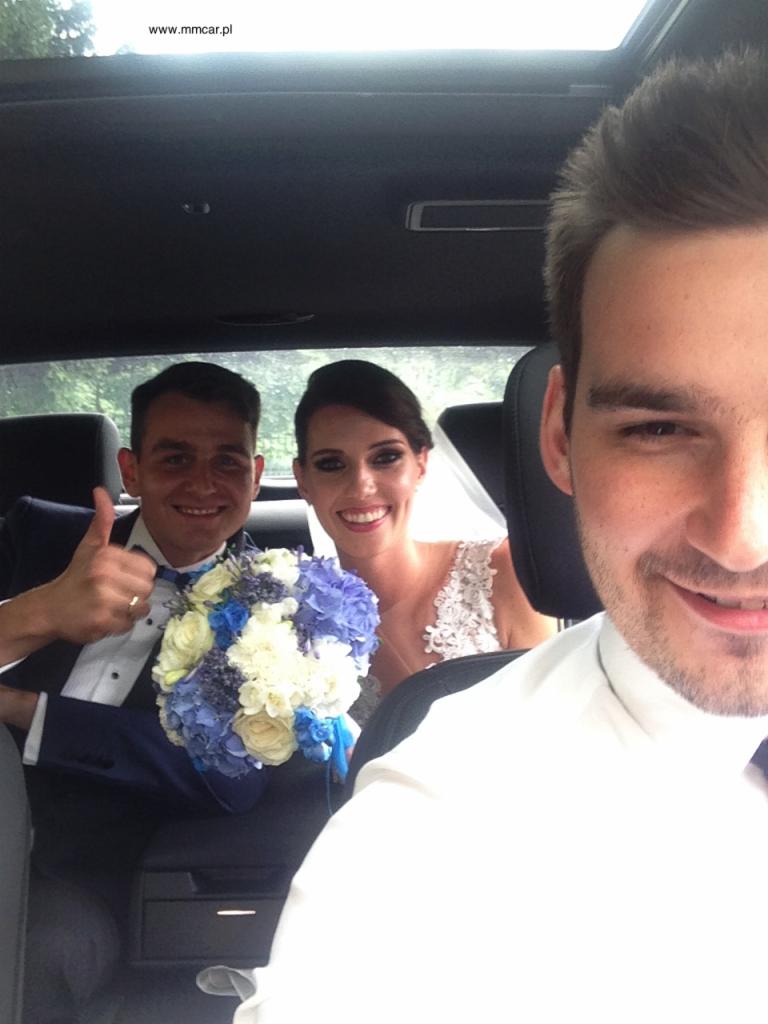 Selfie z wnętrza samochodu: uśmiechnięta para młoda z bukietem kwiatów w odcieniach bieli i błękitu, mężczyzna w garniturze pokazuje kciuk w górę.