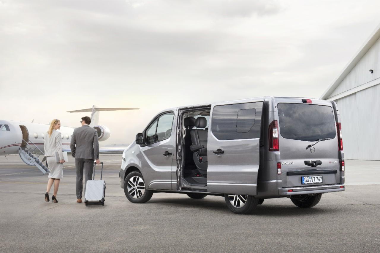 Srebrny van Opel Vivaro z otwartymi drzwiami przesuwnymi stoi na płycie lotniska, para elegancko ubranych ludzi z walizką idzie w kierunku prywatnego odrzutowca w tle.