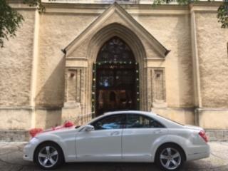 Biały, luksusowy samochód udekorowany czerwonymi kokardami, zaparkowany przed wejściem do zabytkowego kościoła o jasnej fasadzie. Widok z boku, lekko z dołu.