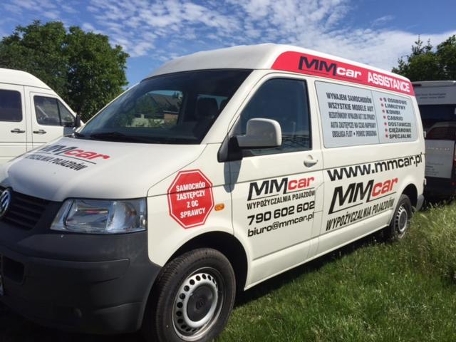 Biały bus z logo MMcar Assistance i numerem telefonu, zaparkowany na trawie. Oferta wynajmu samochodów zastępczych z OC sprawcy. Widoczne inne pojazdy.