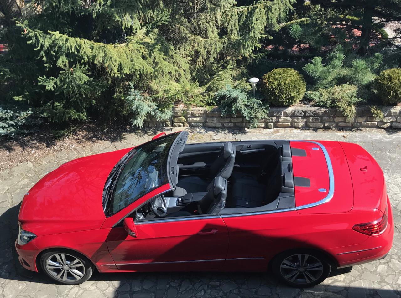 Czerwony kabriolet Mercedes-Benz E-klasy, sfotografowany z góry na tle zielonych drzew i kamiennego muru, z widocznymi czarnymi skórzanymi siedzeniami.