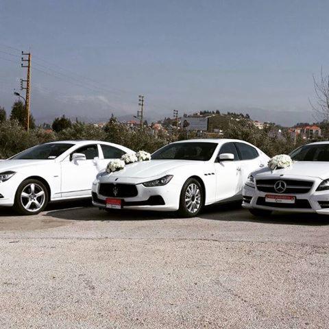 Trzy białe samochody (Mercedes i Maserati) udekorowane białymi kwiatami, ustawione obok siebie na parkingu, prawdopodobnie do ślubu.