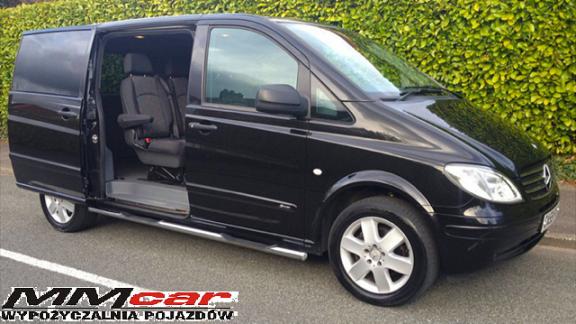 Czarny van Mercedes Vito z otwartymi drzwiami bocznymi, widoczne wnętrze z fotelami, srebrne progi, na tle zielonego żywopłotu, logo wypożyczalni pojazdów w lewym dolnym rogu.