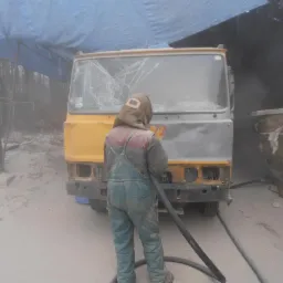 Pracownik w kombinezonie piaskuje uszkodzoną kabinę żółtej ciężarówki z pękniętą szybą przednią, widoczny sprzęt do piaskowania i gruz na zewnątrz.