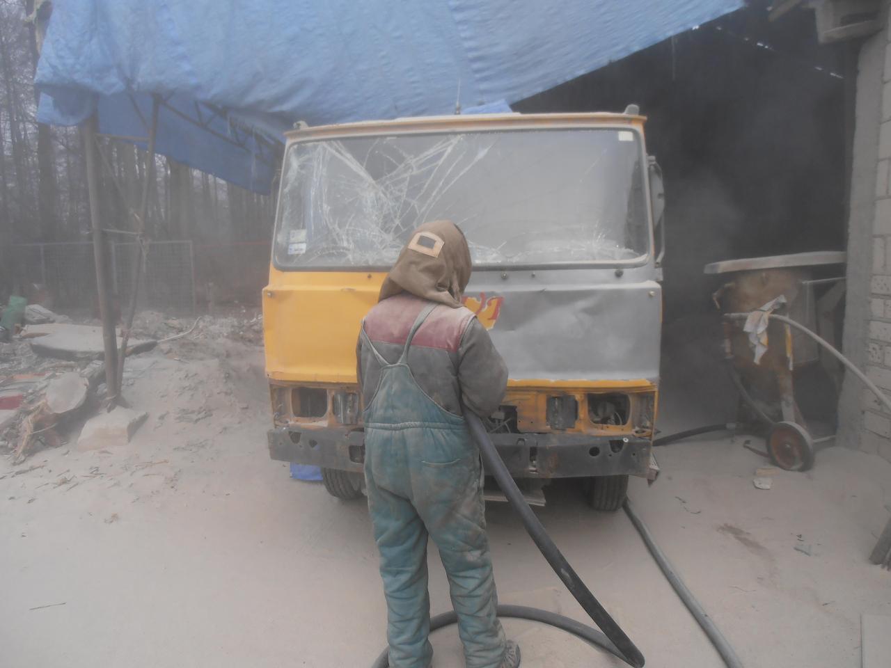 Pracownik w kombinezonie piaskuje uszkodzoną kabinę żółtej ciężarówki z pękniętą szybą przednią, widoczny sprzęt do piaskowania i gruz na zewnątrz.