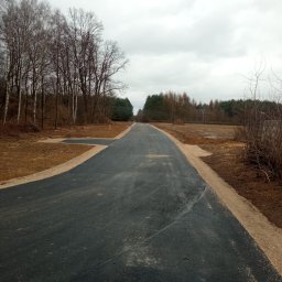 AMK-TRANS - Nowo wyasfaltowana droga przez teren leśny, z widocznymi poboczami z kruszywa i młodymi drzewami po bokach. Pochmurne niebo w tle.