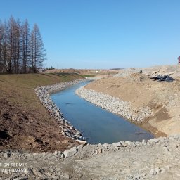 AMK-TRANS - Uregulowany ciek wodny z brzegami umocnionymi kamieniami, w tle ciężarówka i drzewa bez liści, błękitne niebo. Prace ziemne w toku.