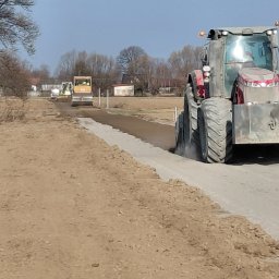AMK-TRANS - Prace drogowe w toku: walec i traktor ubijają nawierzchnię drogi gruntowej. W tle widoczne budynki i drzewa. Słoneczny dzień.
