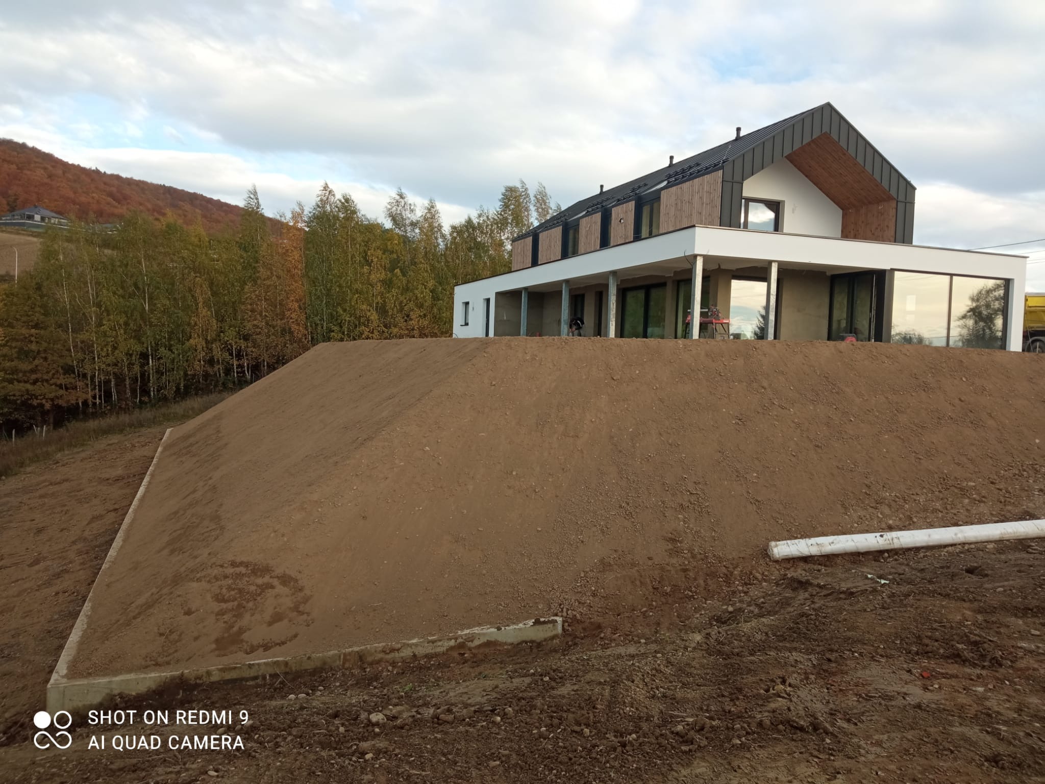 Nowoczesny dom z dużymi oknami i ziemnym nasypem. Widoczne ślady prac ziemnych i rury. W tle drzewa i pochmurne niebo.