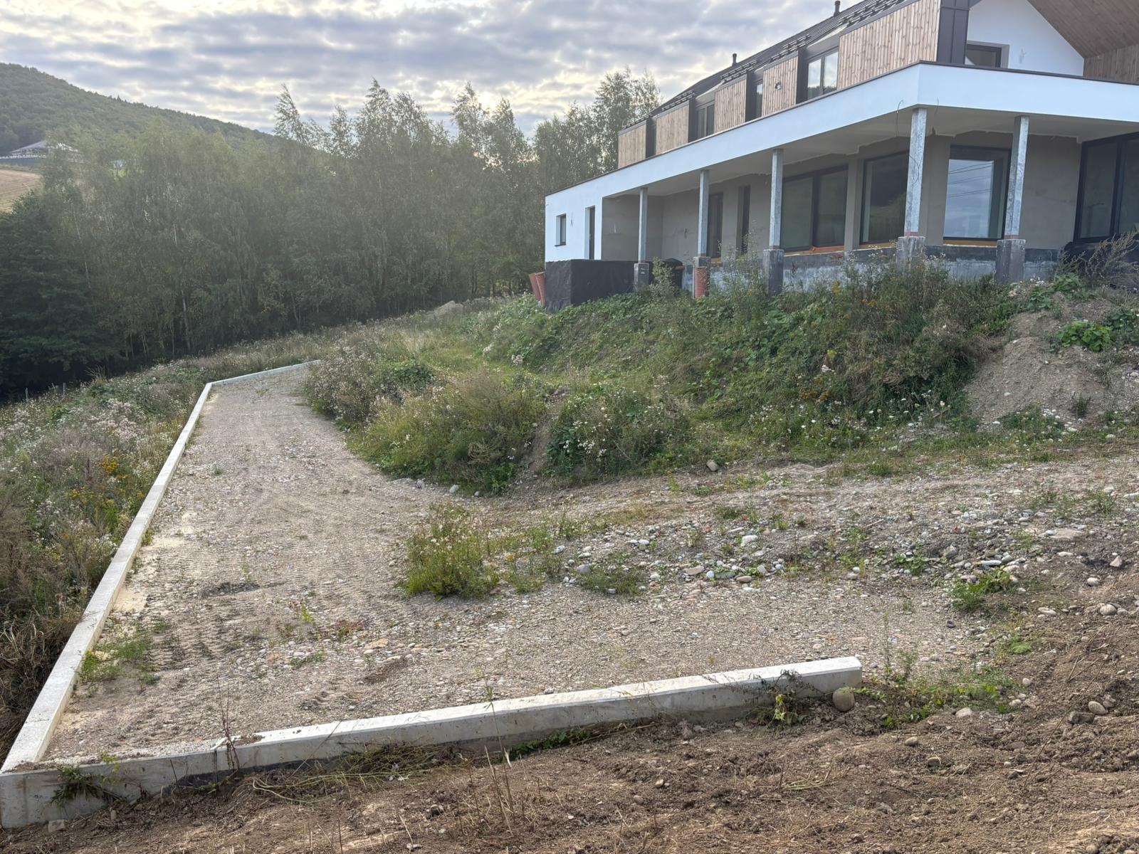Przygotowany teren pod budowę z betonowymi obrzeżami, w tle dom w stanie surowym otoczony zielenią. Widok na zbocze wzgórza i pochmurne niebo.