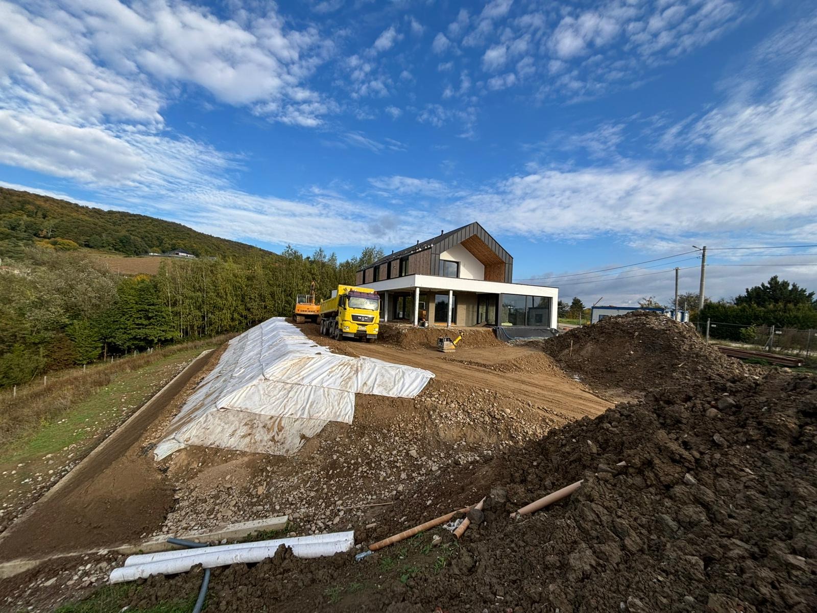 Żółta wywrotka MAN i koparka na placu budowy nowoczesnego domu z dużymi oknami. W tle wzgórze z drzewami i błękitne niebo z chmurami.