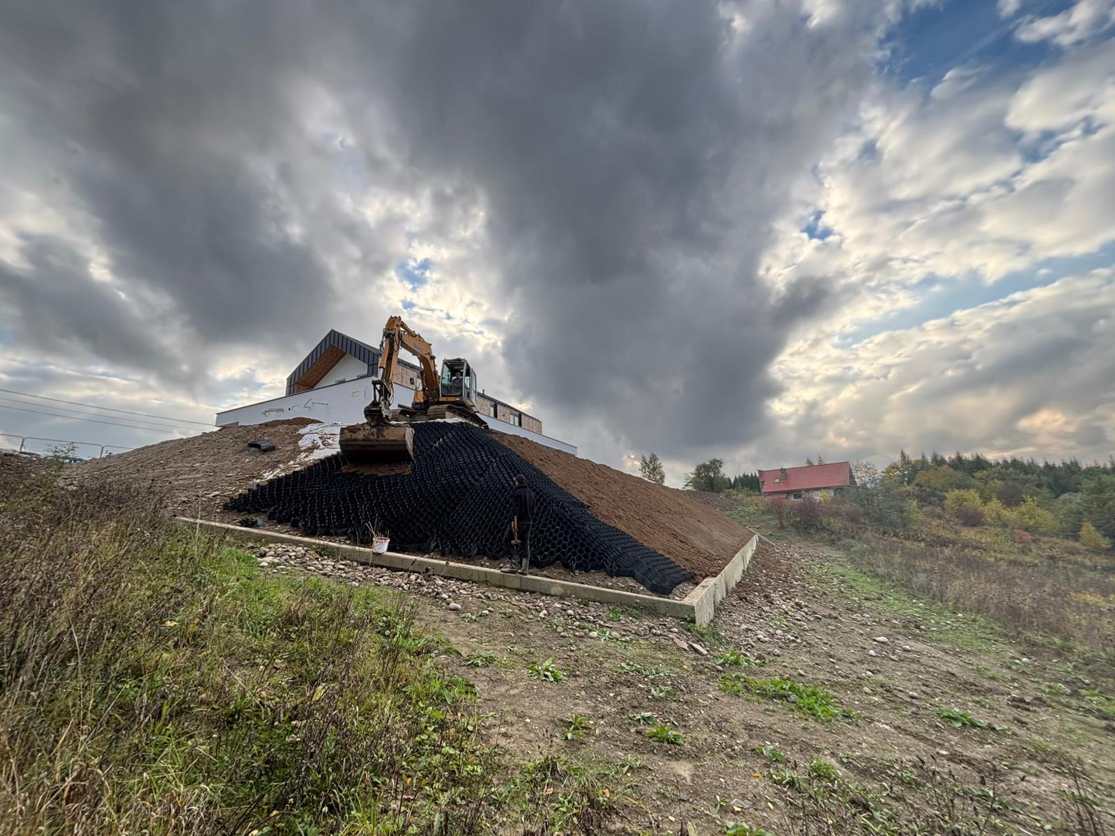 Prace ziemne: koparka układa geokratę na zboczu wzgórza, przygotowując teren pod budowę. W tle widoczny budynek i pochmurne niebo.