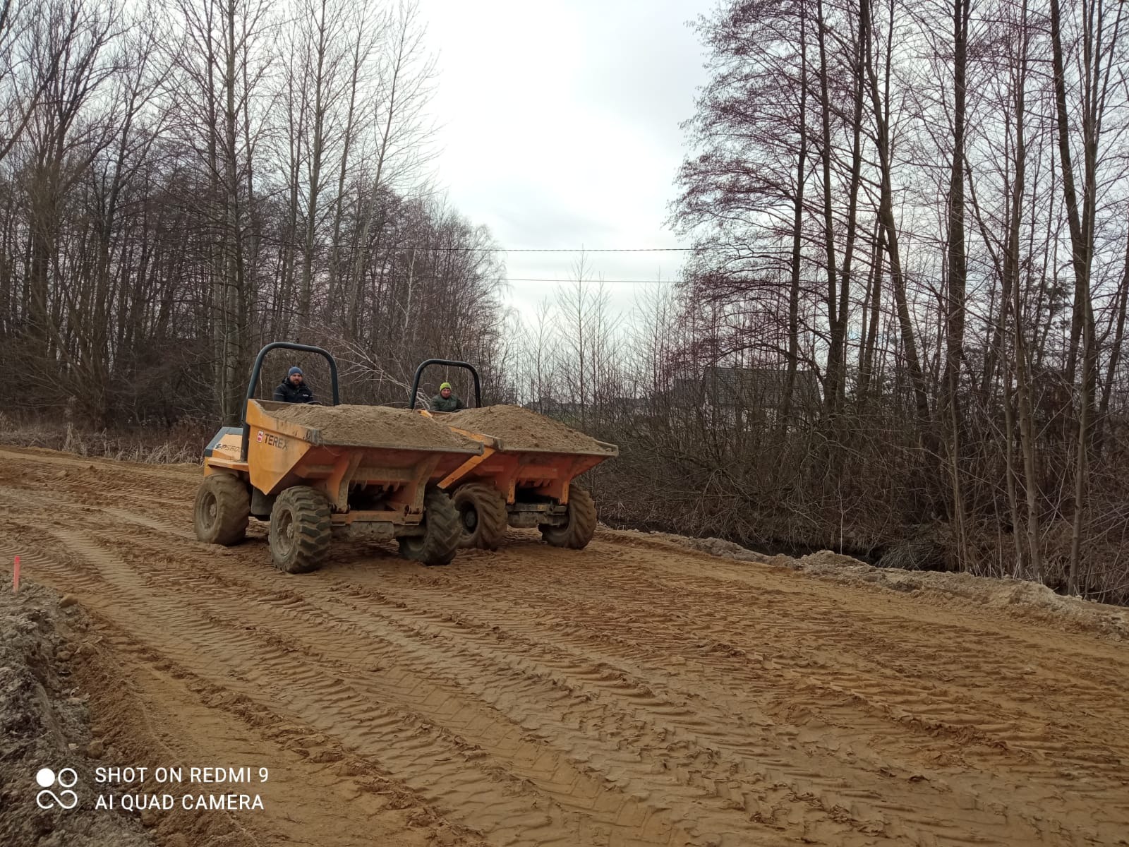 Dwie pomarańczowe wywrotki Terex wypełnione ziemią, jadą po ubitej, piaszczystej drodze. W tle drzewa bez liści i pochmurne niebo. Widoczne ślady opon.