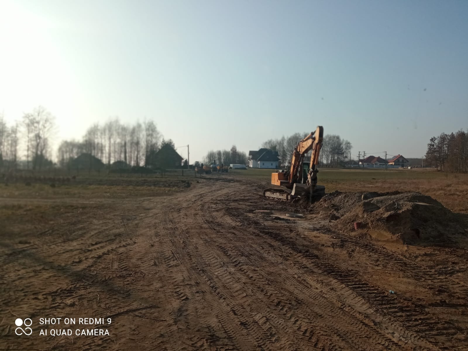 Żółta koparka na placu budowy, świeżo wykopana ziemia, ślady opon na gruncie, w tle zabudowania mieszkalne i drzewa. Słoneczny dzień, błękitne niebo.