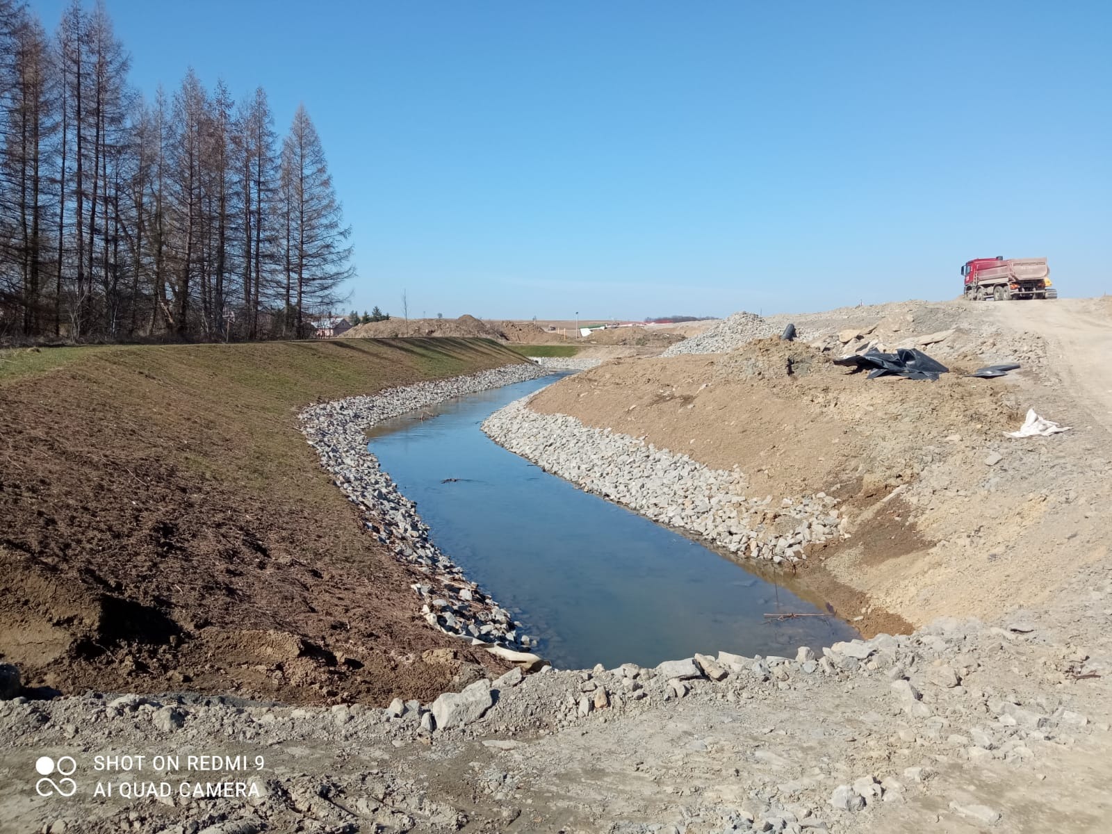 Uregulowany ciek wodny z brzegami umocnionymi kamieniami, w tle ciężarówka i drzewa bez liści, błękitne niebo. Prace ziemne w toku.