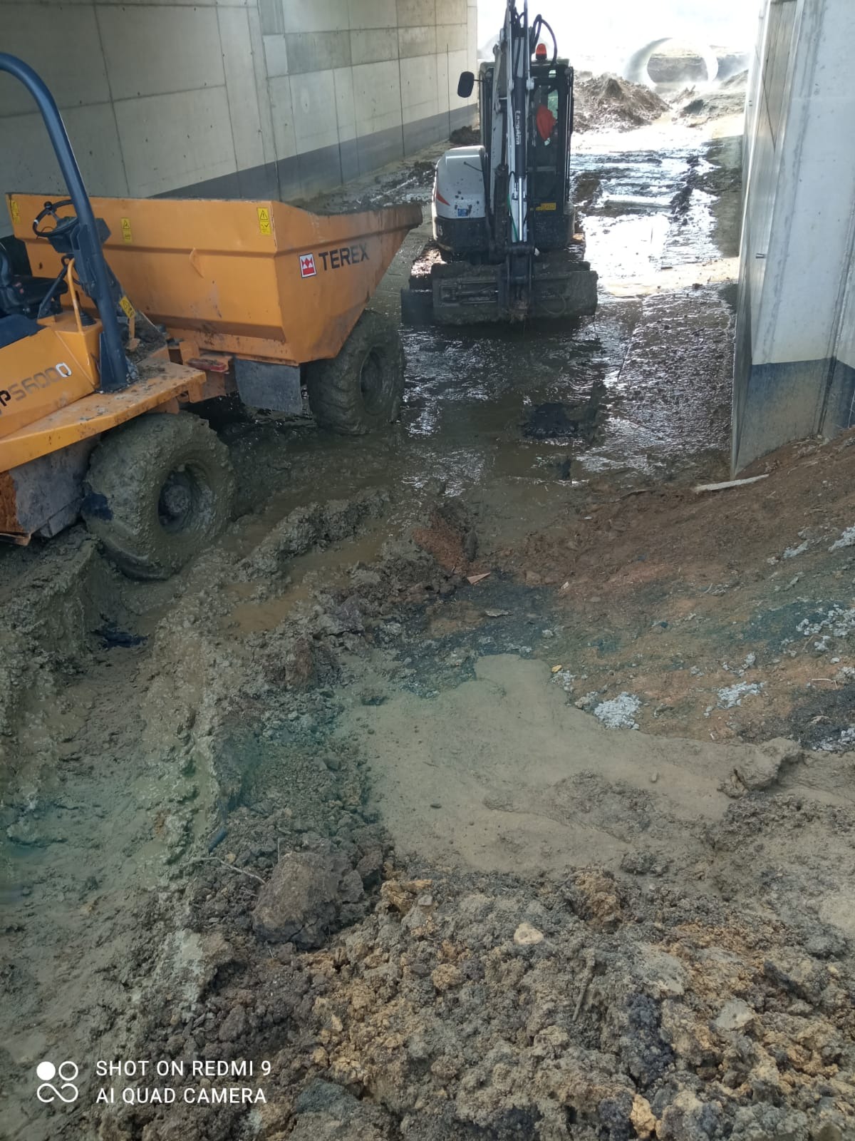 Żółta wywrotka Terex i koparka w błotnistym wykopie pod wiaduktem. Prace ziemne w toku, widoczne ślady opon i świeżo naruszona gleba.
