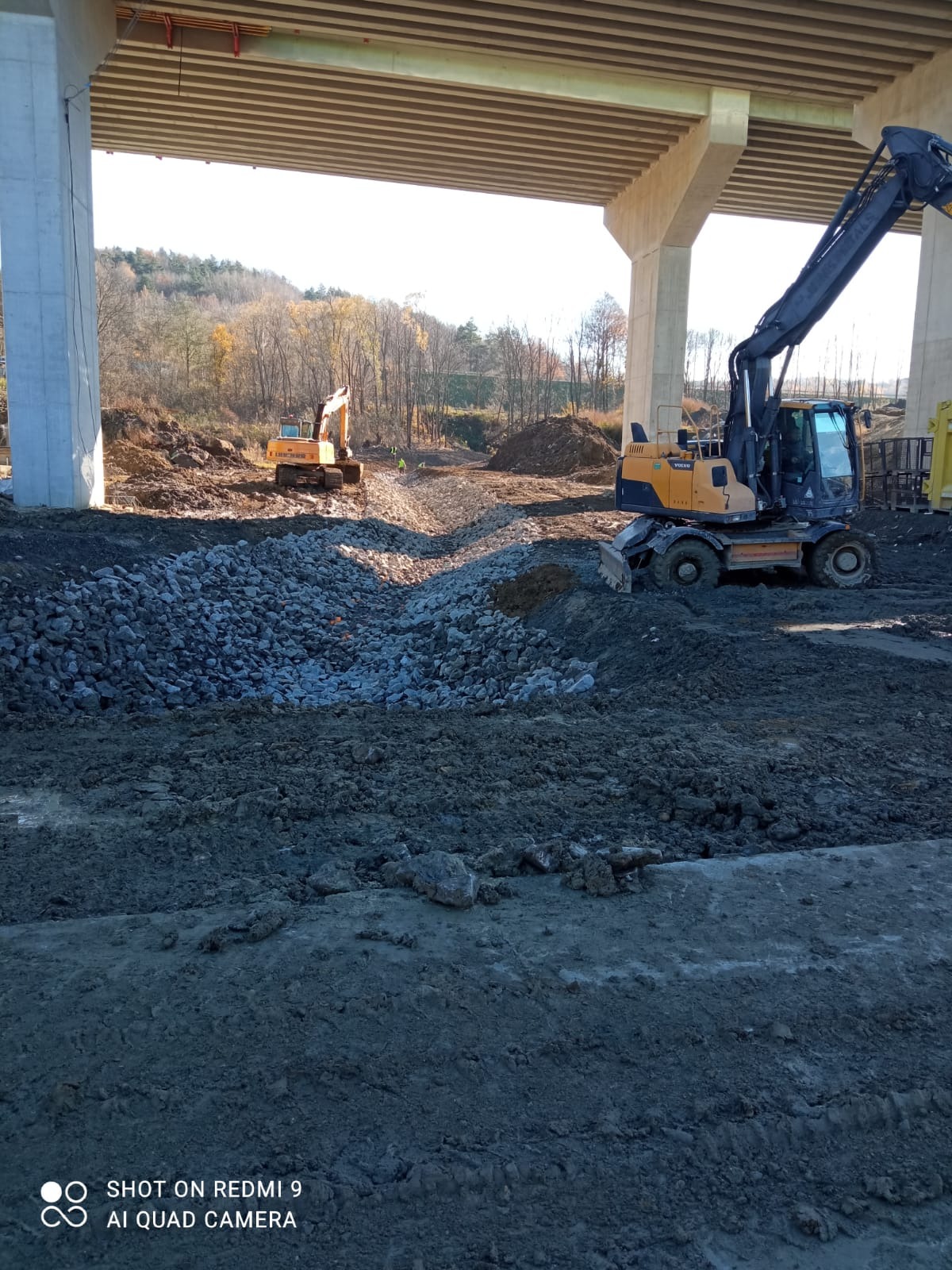 Żółta koparka kołowa Volvo na placu budowy drogi pod wiaduktem, w tle druga koparka i drzewa. Część drogi utwardzona kamieniami.