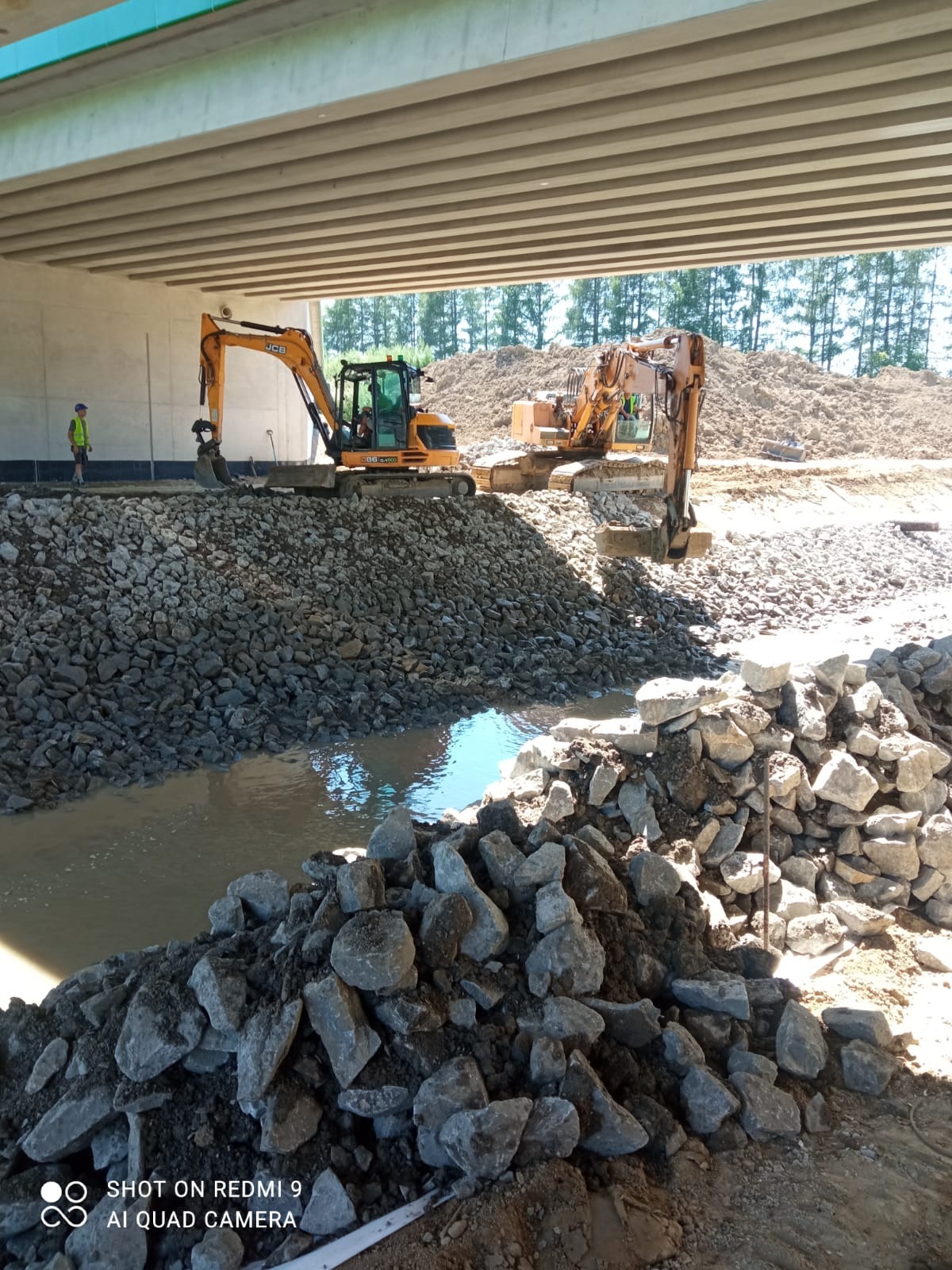 Dwie koparki JCB pracują przy budowie drogi pod wiaduktem, układając kamienie. W tle widać hałdę ziemi i drzewa. Jeden pracownik w odblaskowej kamizelce nadzoruje prace.