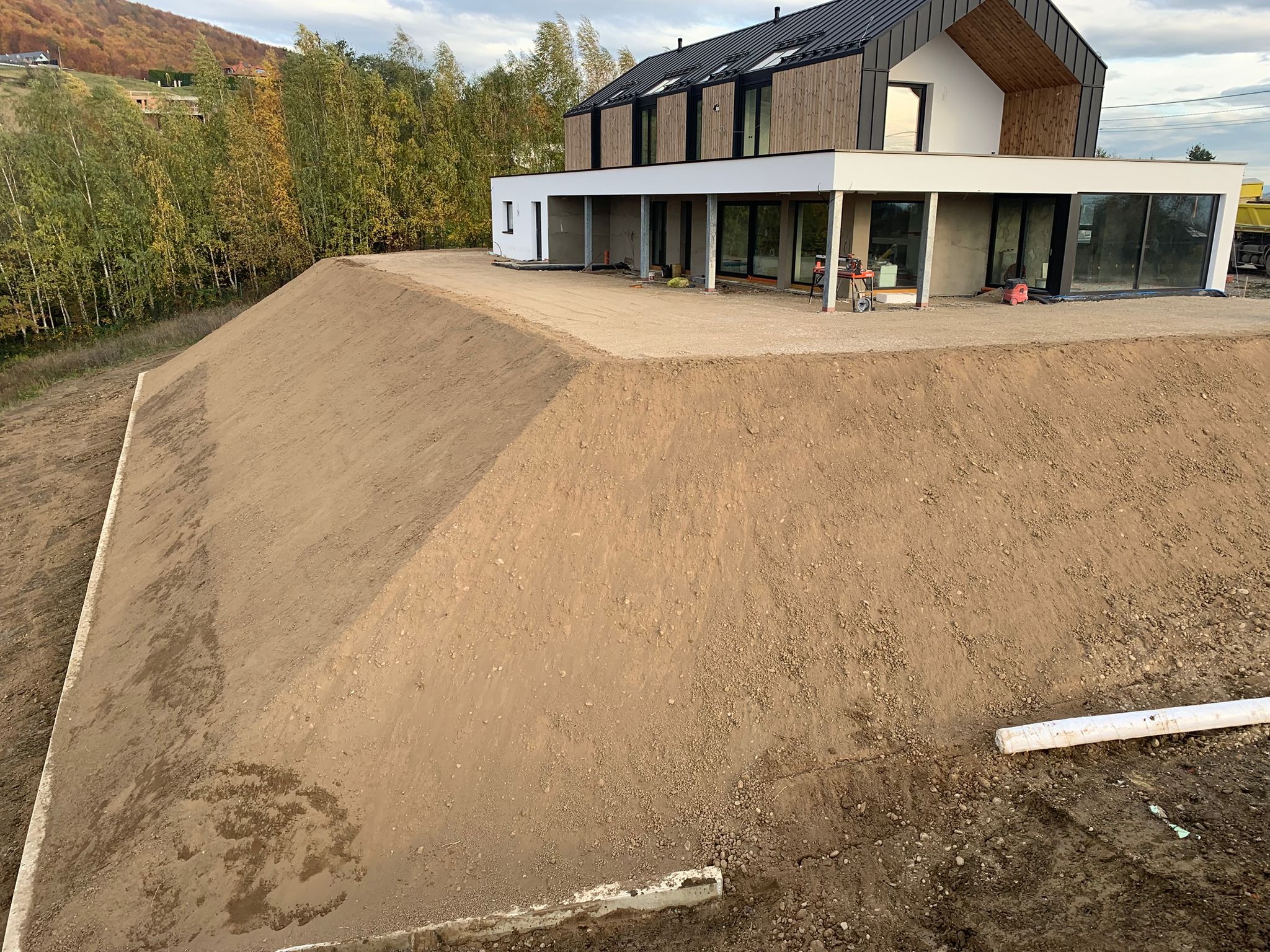 Niwelacja terenu pod nowoczesny dom z drewnianą elewacją. Wyrównany teren z widoczną skarpą i elementami konstrukcyjnymi. Tło stanowią drzewa i wzgórza.