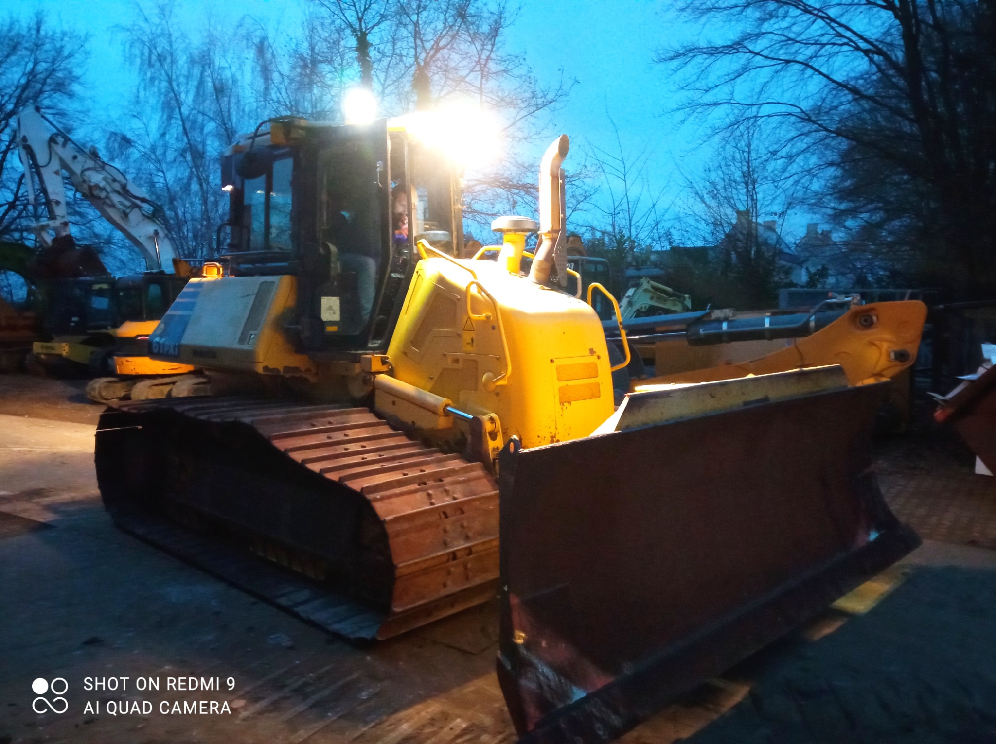 Żółty spychacz gąsienicowy z włączonymi światłami roboczymi, zaparkowany na placu budowy o zmierzchu. W tle widoczne inne maszyny budowlane i drzewa.