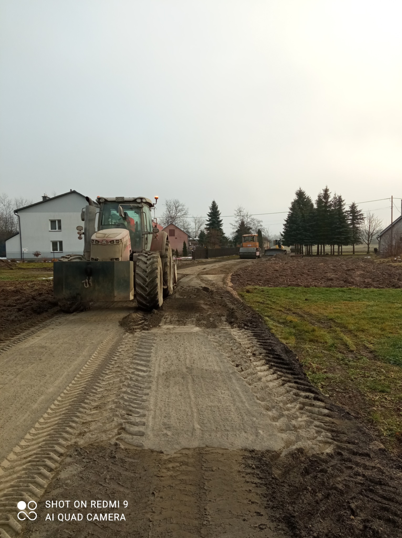 Ciężki traktor z przodu, walec i koparka w tle, na drodze gruntowej z wyraźnymi śladami opon. Prace ziemne w toku, widok na wiejski krajobraz w pochmurny dzień.