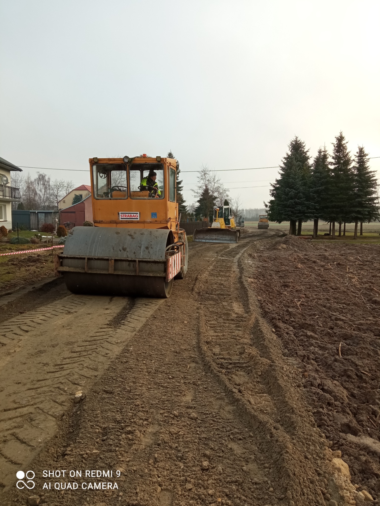 Walec drogowy STRABAG ubija grunt na budowie drogi. W tle widoczny spychacz i drzewa iglaste. Ślady opon na ziemi. Praca w toku, etap utwardzania podłoża.