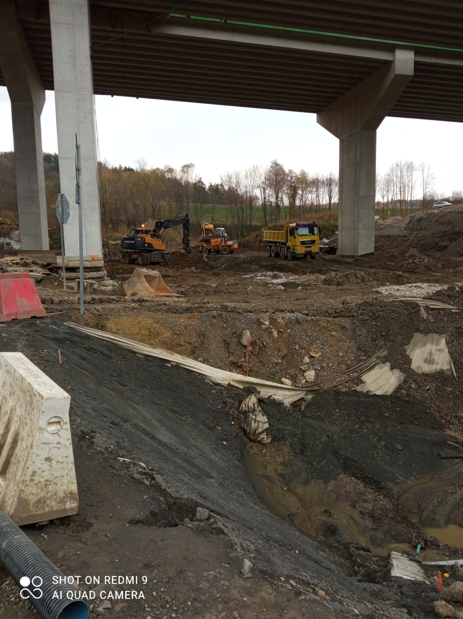 Prace ziemne pod estakadą: koparki Volvo i MAN w akcji. Żółta wywrotka czeka na załadunek. Widoczne zabezpieczenia wykopu i rury drenażowe. Pochmurne niebo w tle.