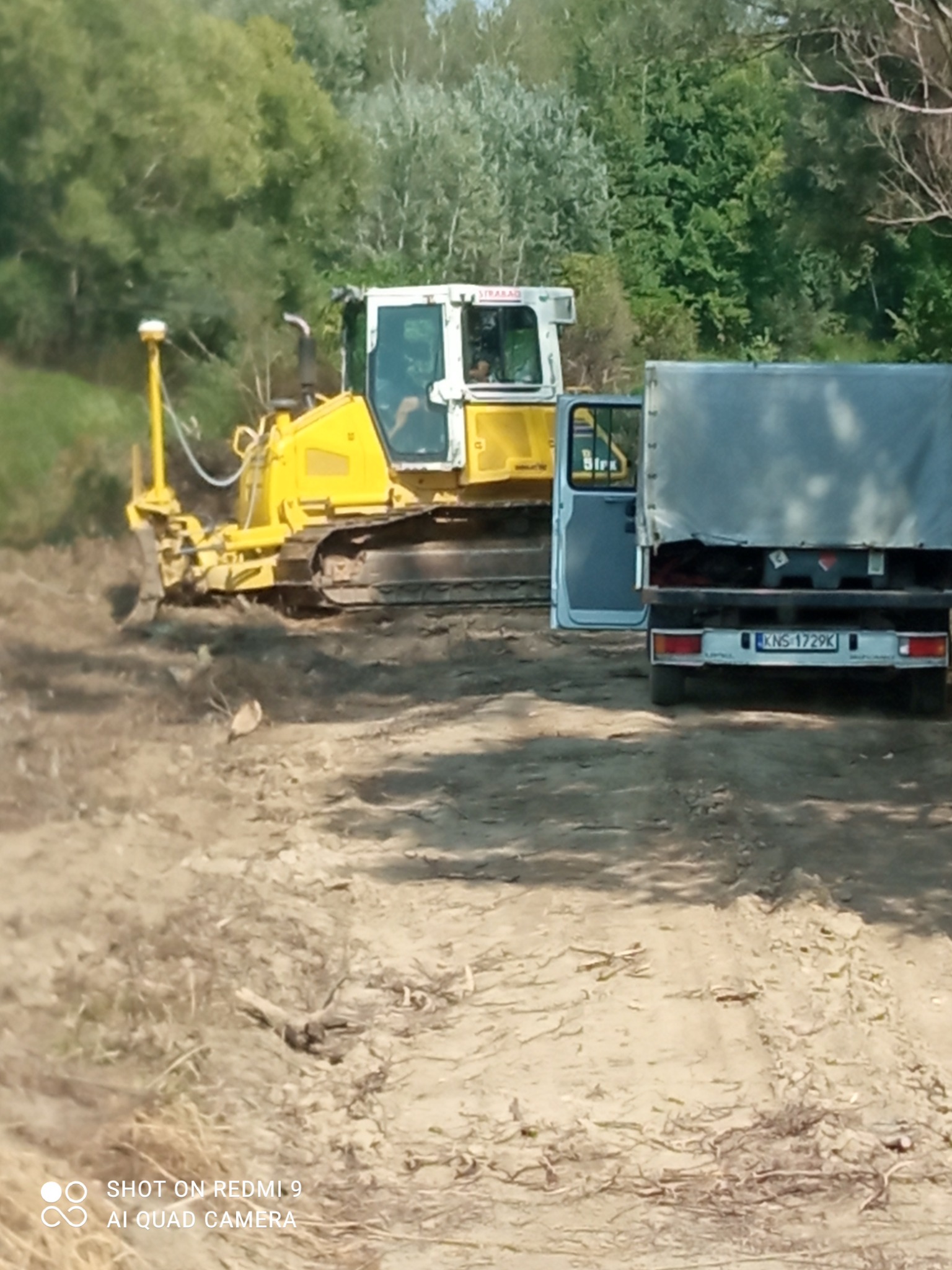 Żółty spychacz Komatsu z GPS na nierównym terenie obok otwartych drzwi ciężarówki. W tle drzewa i krzewy. Widoczne ślady pracy maszyny.